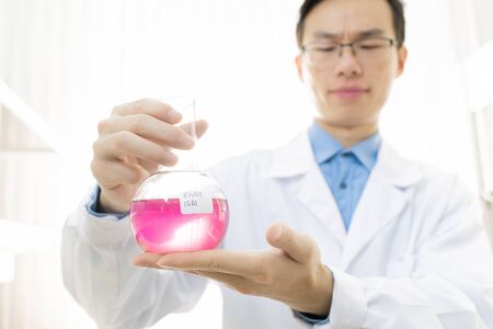 Horizontal portrait of young medical scientist wearing white coat and eyeglasses holding flask with...
