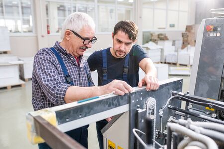 Senior worker in glasses explaining printing machine setups to young colleague at modern factory