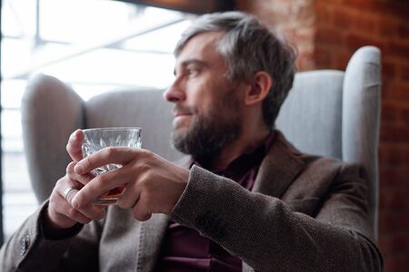 Close-up of introspective middle-aged man in jacket sitting in armchair in living room and enjoying hard liquorの写真素材