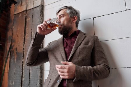 Below view of thoughtful stylish man with beard standing at wall and drinking whisky
