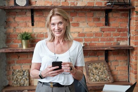 Portrait of smiling modern mature female manager with blond hair surfing net on smartphone
