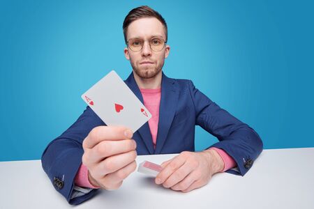 Portrait of serious young gambler in jacket sitting at table against blue background and showing ace...