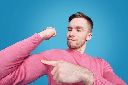 Content confident young bearded man in pink sweater flexing bicep against blue background