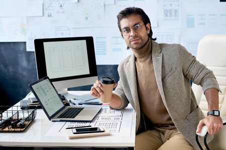 Stylish IT specialist sitting relaxed at office desk holding cup of coffee looking at camera,...