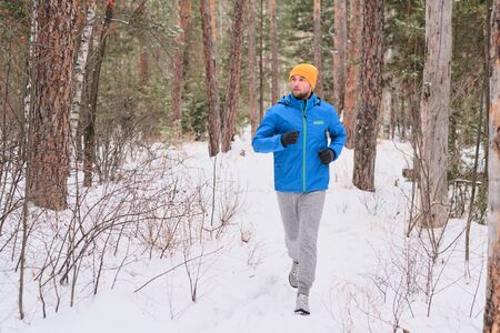 Handsome young man in hat running on snowy path in beautiful forest while training aloneの写真素材
