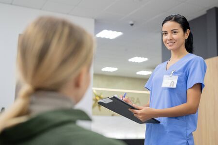 Pretty young brunette assistant in blue uniform looking at one of patients