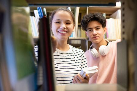 Cheerful teenage girl and her classmate standing by bookshelf in college libraryの写真素材