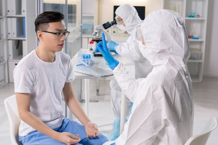 Medical specialist in protective suit showing finger to infected Asian patient while checking his...