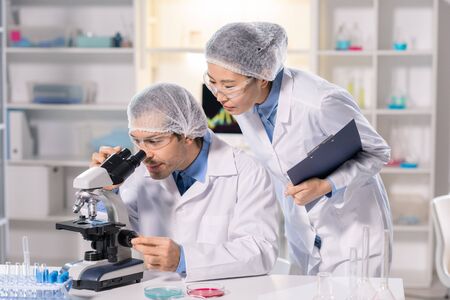 Focused scientist in safety goggles using microscope while working on medical research with Asian...