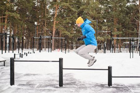 Side view of young man in blue jacket training using horizontal bars as obstacles for jumping in...