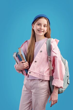Portrait of smiling pretty schoolgirl with satchel holding heap of books against blue backgroundの写真素材