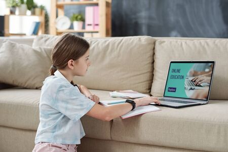 Teenage girl with ponytail sitting at sofa and using laptop while taking online educational course...