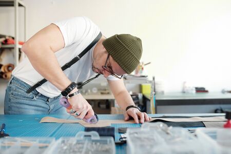 Young craftsman in casualwear and beanie bending over table while cutting piece of beige suede or leather in workshopの写真素材