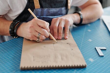Hands of contemporary craftsman with pencil over notepad making sketch of leather item while sitting...