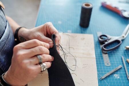 Hands of leatherworker with piece of black leather sewing new item while holding it over table with suppliesの写真素材