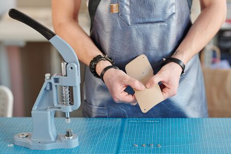 Hands of creative leatherworker in black leather apron putting tiny metallic knobs into light beige...