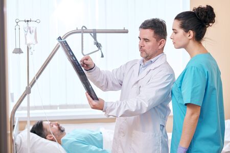 Serious mature doctor in lab coat pointing at x-ray image while discussing results with nurse in...