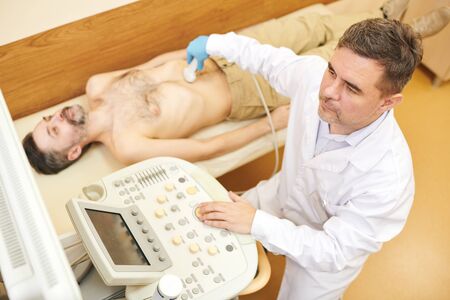 Above view of concentrated sonographer in lab coat using transducer probe on belly of patient during...