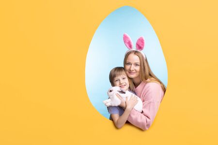 Portrait of happy young mother in hare ears headband hugging bunny with son, egg-shaped frame