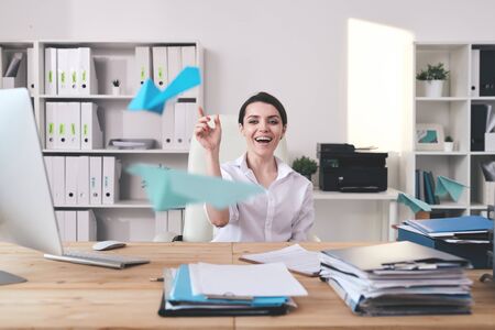 Cheerful excited young businesswoman sitting at desk full of documents and throwing paper plane in officeの写真素材