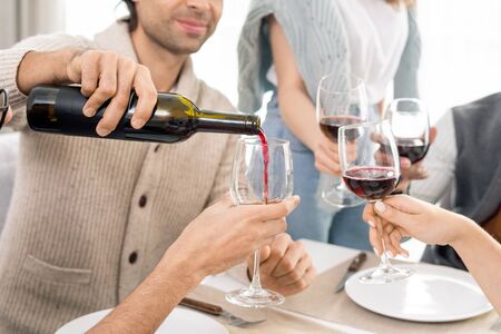 Young man pouring red wine from bottle into wineglasses of his friends while celebrating holiday by...