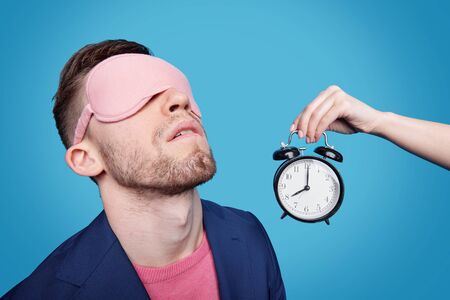 Female hand holding alarm clock close to young man with sleeping mask on his eyes while napping against blue backgroundの写真素材