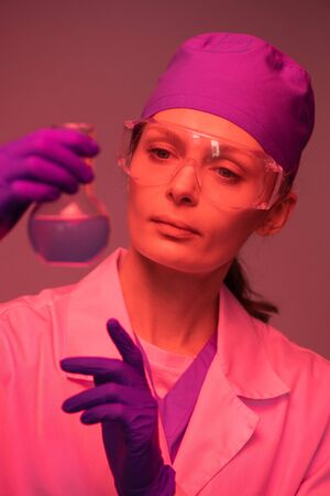 Female scientist in cap and safety goggles working with chemicals in laboratory, red light effectの写真素材