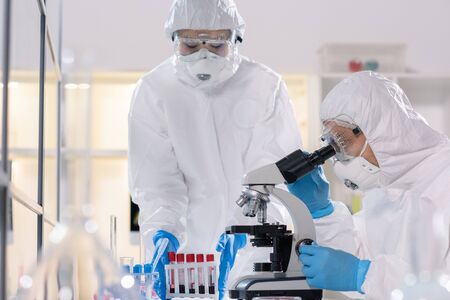 Young scientist in protective workwear looking in microscope while studying new virus with colleague...