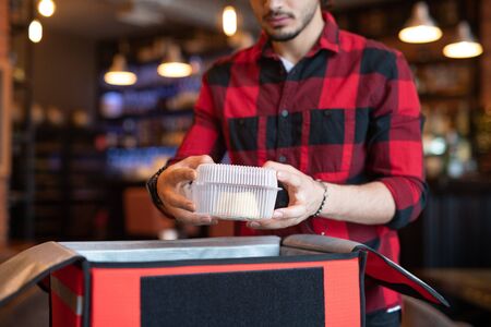 Young courier of cafe putting container with food into big bag
