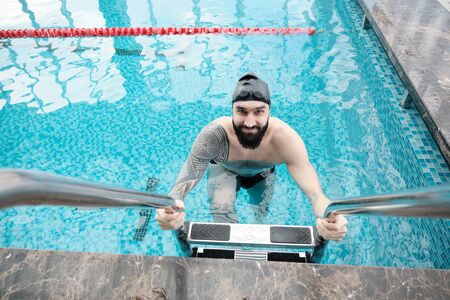 Portrait of smiling young bearded swimmer in black cap getting out of water using stairs in pool