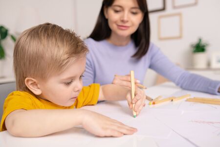 Close-up of concentrated cute boy sitting at table and using pencils for drawing under control of motherの写真素材
