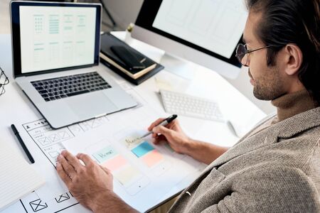 Serious phone interface engineer in glasses sitting at desk and making notes on interface sketch