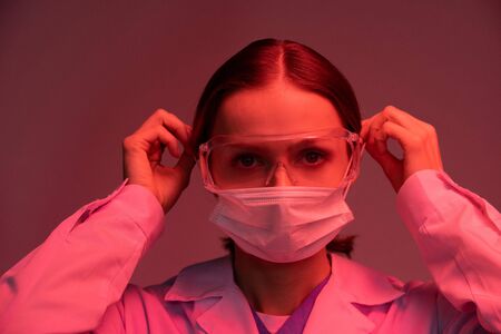 Virologist in protective eyeglasses and whitecoat putting mask on her face