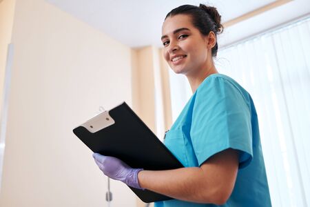 Portrait of smiling attractive nurse in surgical gloves making notes in medical card while doing rounds in hospitalの写真素材