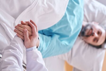 Close-up of doctor holding hands of mature patient in bed while supporting him in hospitalの写真素材