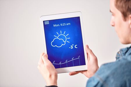Over shoulder view of young man checking weather forecast on tablet app against white wallの写真素材