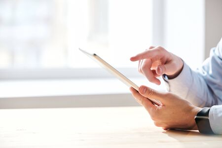 Close-up of unrecognizable businessman sitting at table and using tablet while checking business...
