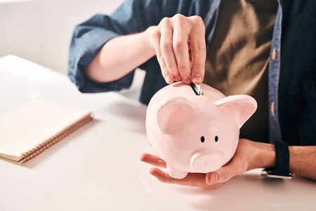 Close-up of unrecognizable man sitting at table and putting coins into piggybank while saving money in crisisの写真素材