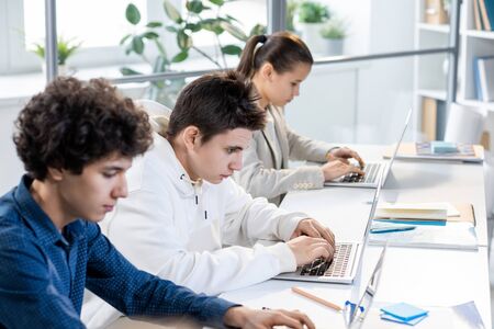 Group of three teenage students sitting in row by desk and working individually