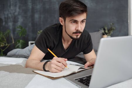 Concentrated young bearded student lying in bed and making notes while listening to lecture online...