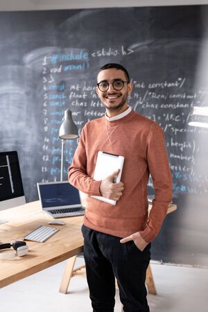 Portrait of cheerful young middle-eastern IT specialist in glasses standing with tablet in modern...