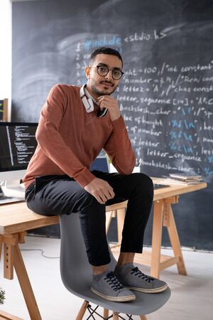 Portrait of serious young Arabian IT engineer sitting on table with feet on chair in modern office...