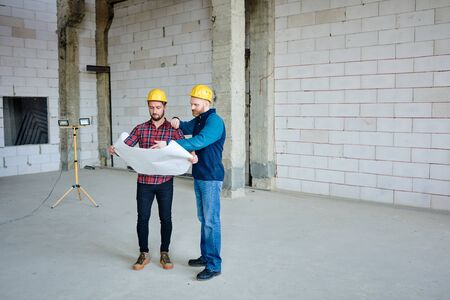 Two young builders in protective helmets discussing sketch in blueprintの写真素材