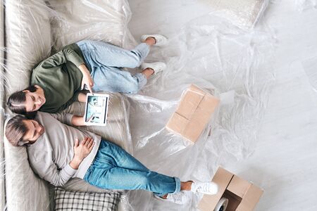 Overview of young affectionate couple with tablet sitting on new couch