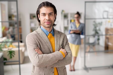 Portrait of content young business leader in beige jacket standing with crossed arms in modern...