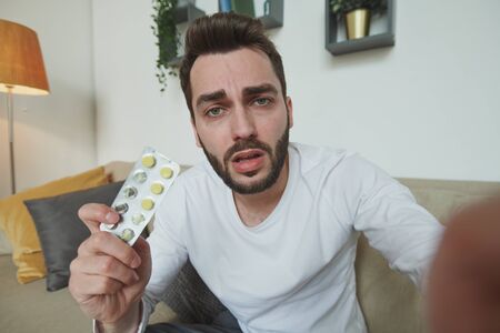 Bearded man with cold or covid19 showing blister with tablets to online doctorの写真素材