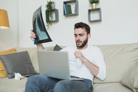 Young bearded man with lungs x-ray result sitting on couch in front of laptop