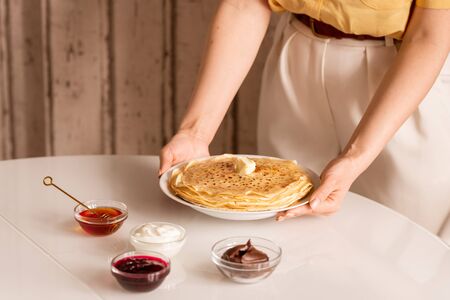 Hands of young housewife putting plate with stack of hot homemade pancakes on table with honey, sourcream, jam and chocolateの写真素材