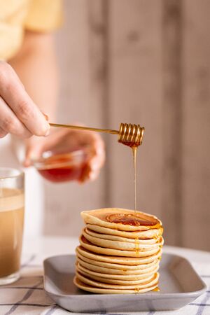 Hand of young housewife standing by table and putting fresh honey on top of pancake stack while cooking breakfast for her family in the kitchenの写真素材