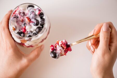 Overview of female hands holding glass and spoon with homemade yoghurt consisting of muesli, blackberries or black currant, jam and sourcreamの写真素材